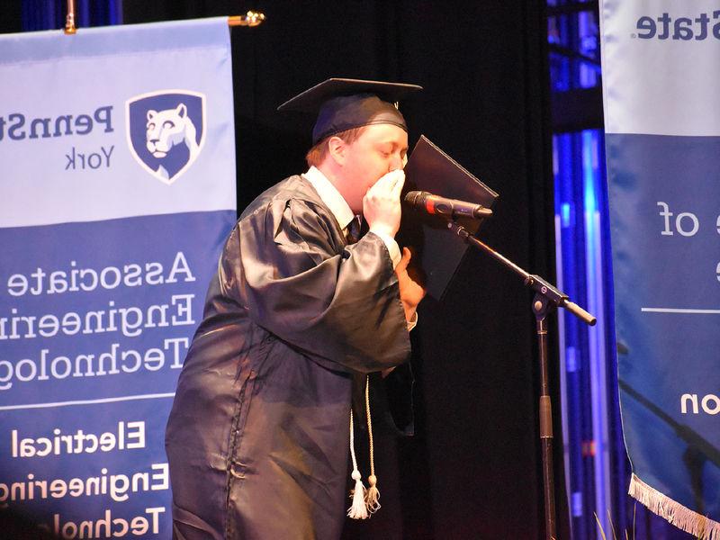 Male student in graduation cap and gown speaking into a microphone on a stabnd